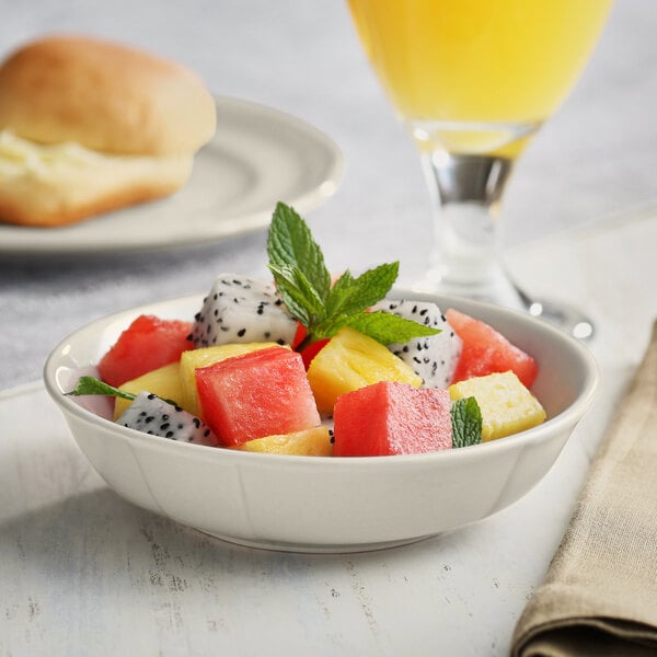 A bowl of fruit salad in an Acopa Warm Gray Scalloped Porcelain fruit dish on a table next to a glass of orange juice.