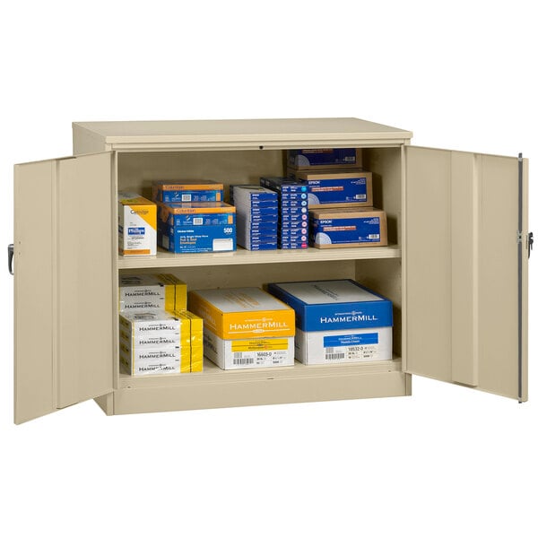 A sand Tennsco storage cabinet with solid doors open to reveal boxes on shelves.