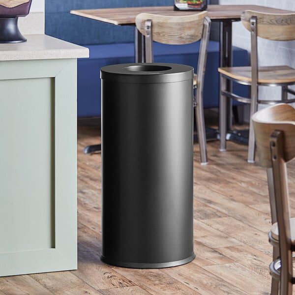 A black Lancaster Table & Seating decorative waste receptacle on a wood floor.