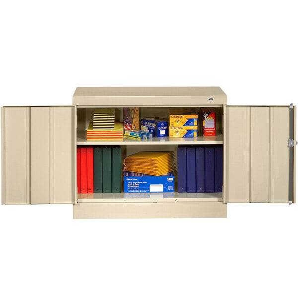 A sand Tennsco standard storage cabinet with solid doors filled with books and folders.