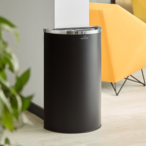 A black Lancaster Table & Seating half round decorative trash can next to a yellow chair.