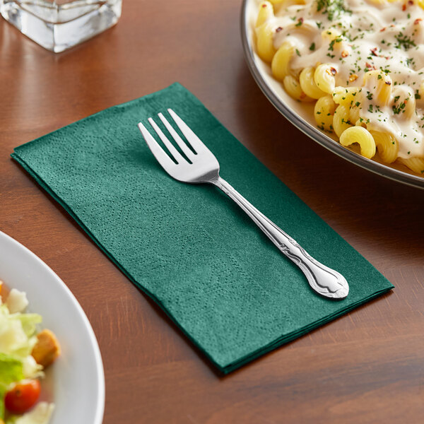 A fork on a green Hoffmaster paper napkin next to a plate of pasta and salad.