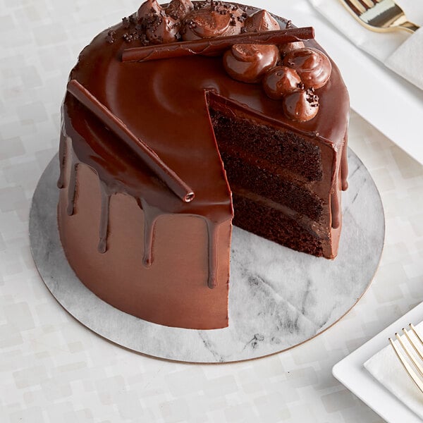 An Enjay round cardboard marble and wood reversible cake board with a chocolate cake on it in a bakery display with a slice missing.