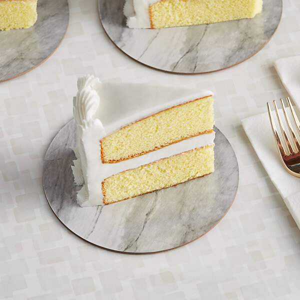 A slice of yellow cake with white frosting on an Enjay marble and wood laminated cake board with a fork.