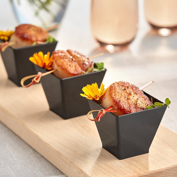 A group of small black Choice square plastic shot glasses filled with food on a table.