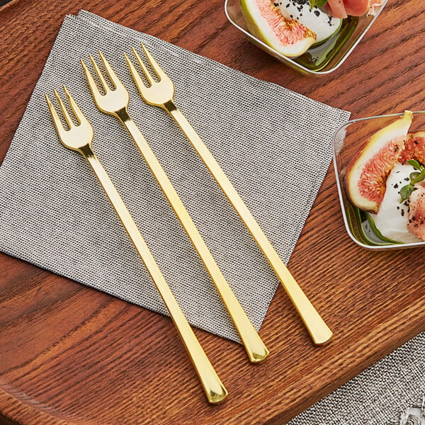 A group of Visions gold plastic tasting forks on a table next to a bowl of fruit.