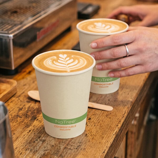 Two cups of latte with leaf-shaped latte art in compostable paper cups are placed on a wooden counter, with one cup being picked up by a hand.