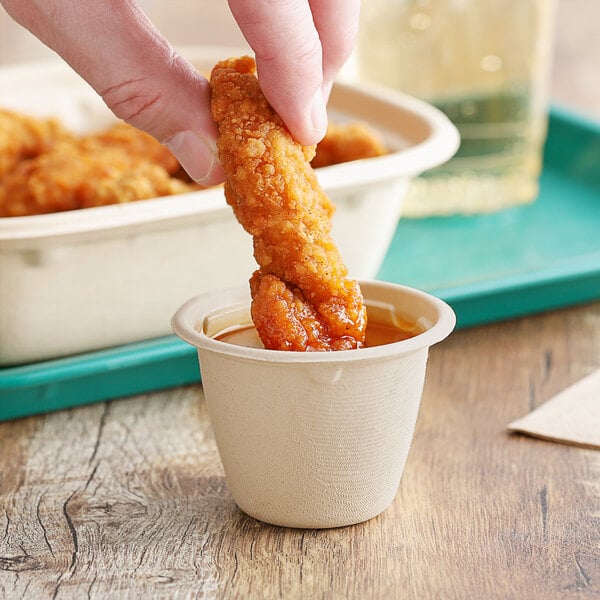 A person holding a World Centric compostable fiber portion cup with a piece of fried chicken dipped in sauce.