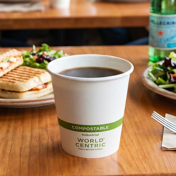 A white compostable paper cup filled with a dark beverage is placed on a wooden table, with plates of food and a bottle in the background.