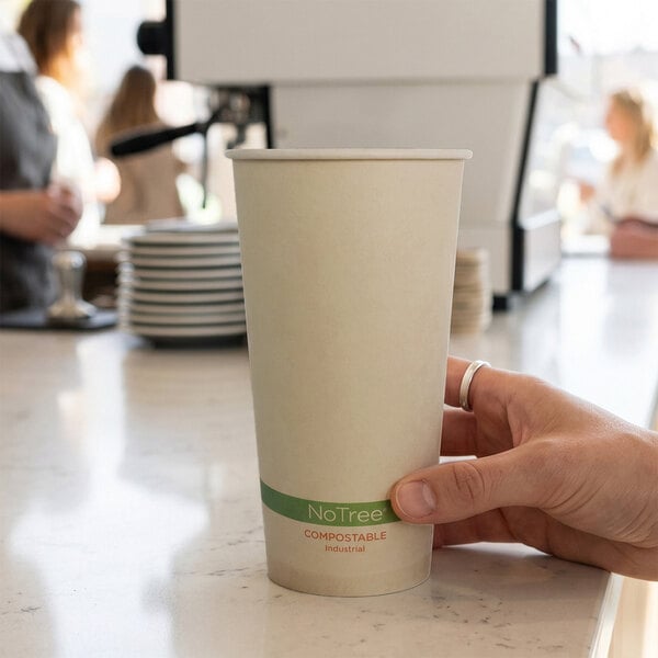 A hand is holding a plain, tall, compostable paper cup with a green band near the bottom, on a countertop in a café setting.