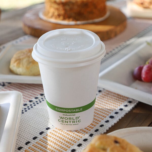 A white disposable cup with a lid and a green band labeled 'COMPOSTABLE' sits on a table among plates of food.