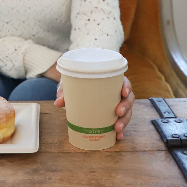 A person is holding a beige paper cup with a white lid and a green band labeled 'NoTree COMPOSTABLE' while sitting at a wooden table next to a plate with a pastry.