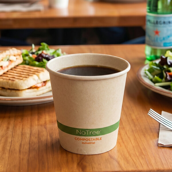 A compostable paper cup filled with a dark beverage is placed on a wooden table, with plates of food and a bottle in the background.