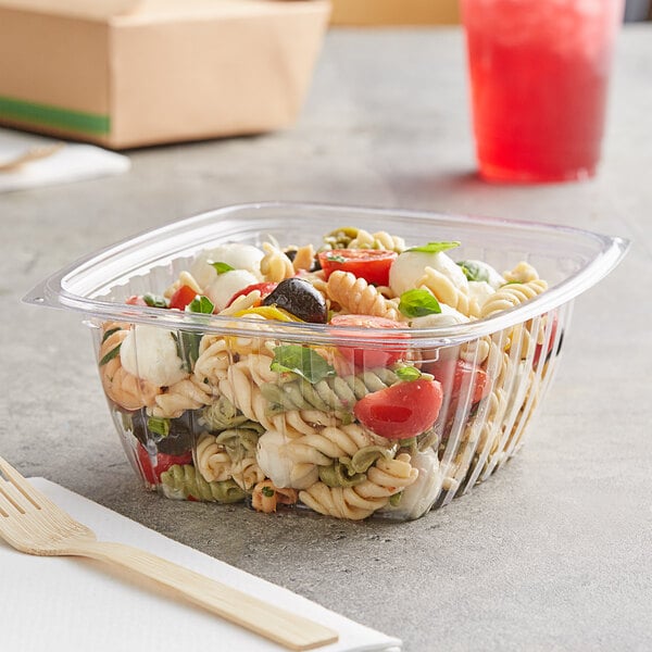 A World Centric clear plastic deli container filled with pasta and tomatoes on a table with a wooden spoon.