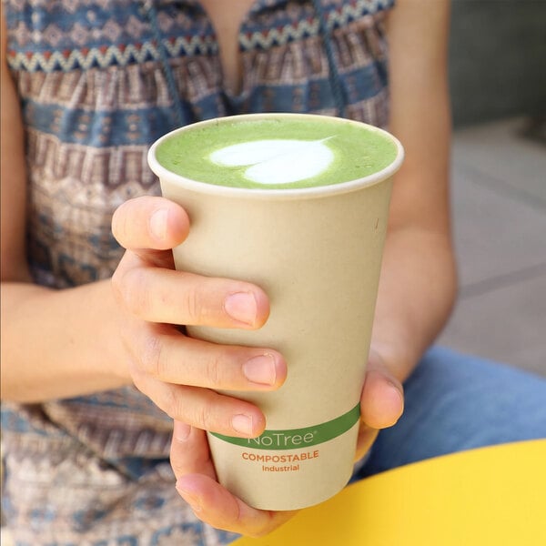 A person is holding a compostable paper cup filled with a green beverage topped with a heart-shaped foam design.