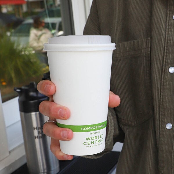 A person is holding a white compostable paper cup with a lid and a green label that reads 'COMPOSTABLE' and 'WORLD CENTRIC.'