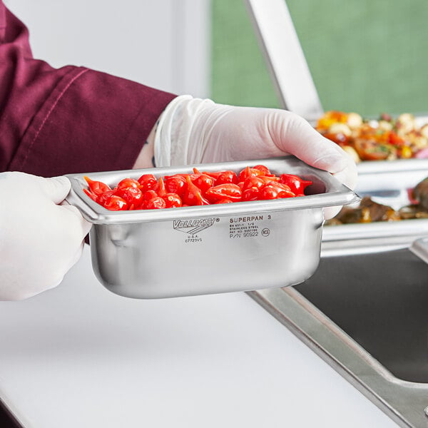 A person in gloves holding a Vollrath stainless steel container of tomatoes.