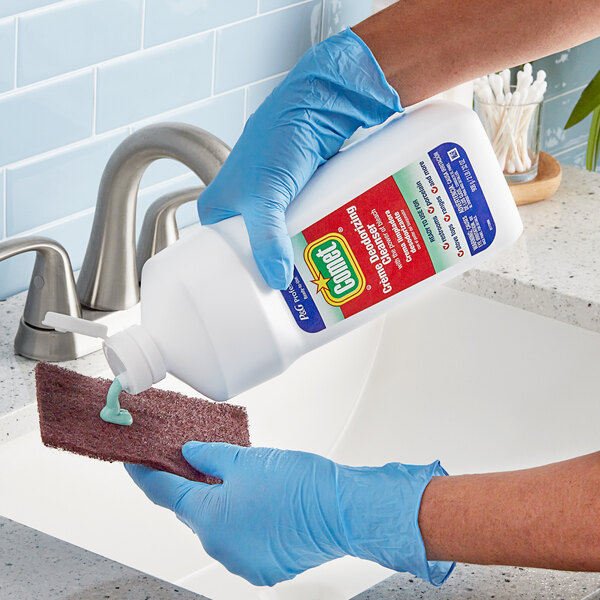 A person in blue gloves pouring Comet Creme Deodorizing Cleanser into a sponge to clean a sink.