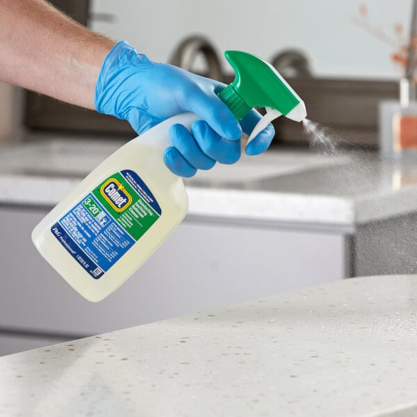 A hand in blue gloves holding a Comet spray bottle and spraying a white counter.