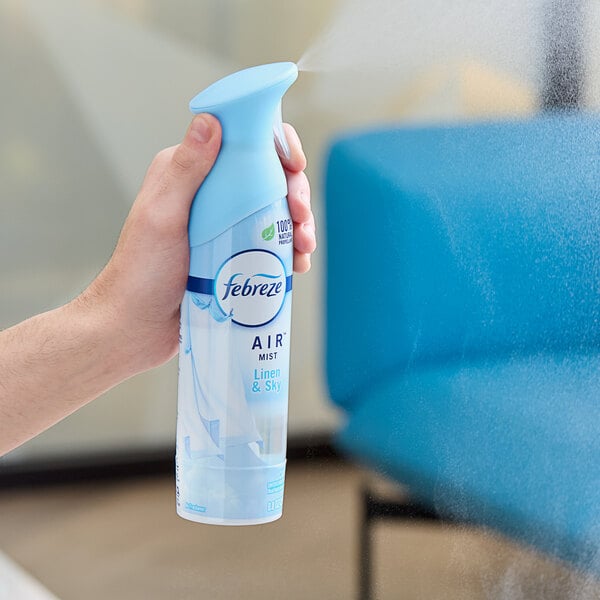 A hand spraying Febreze Air Linen & Sky scented air freshener in a white room.