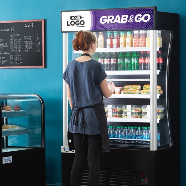 A woman standing in front of an Avantco black refrigerated air curtain merchandiser.