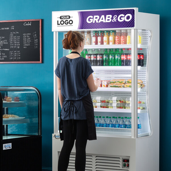 An Avantco white refrigerated air curtain merchandiser with a woman in a black apron standing in front.