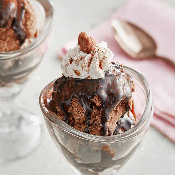 A glass cup of chocolate ice cream with whipped cream and a spoon with a jar of Oringer Premium Fudge Dessert on the side.