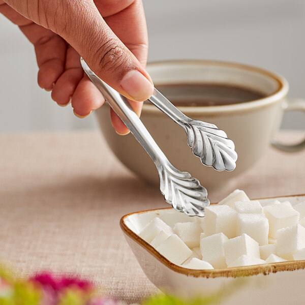 A person using American Metalcraft stainless steel sugar tongs to pick up sugar cubes from a bowl.