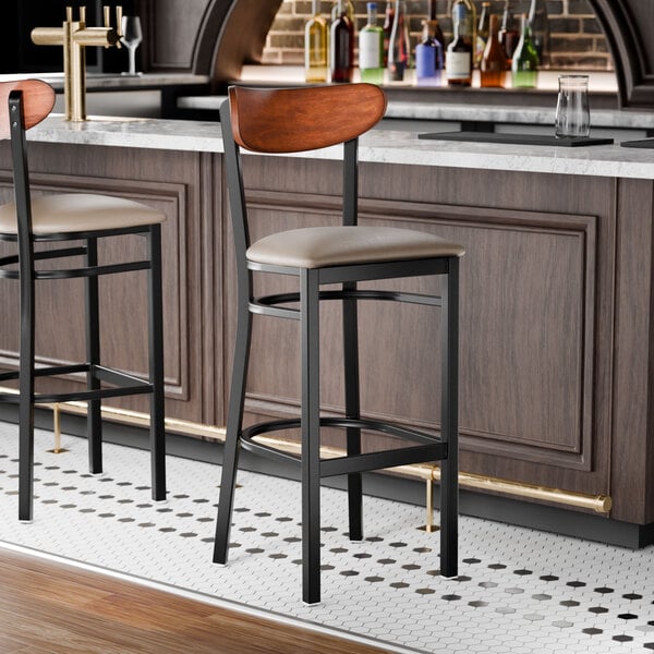 A bar stool with a black metal frame, taupe vinyl seat, and an antique walnut wood backrest.