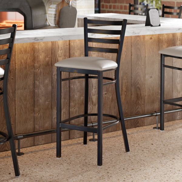 A ladder back bar stool with a distressed copper finish and a light gray vinyl padded seat.