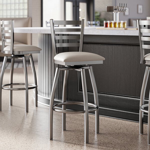 A group of Lancaster Table & Seating clear coat finish ladder back swivel bar stools with light gray padded seats at a bar.