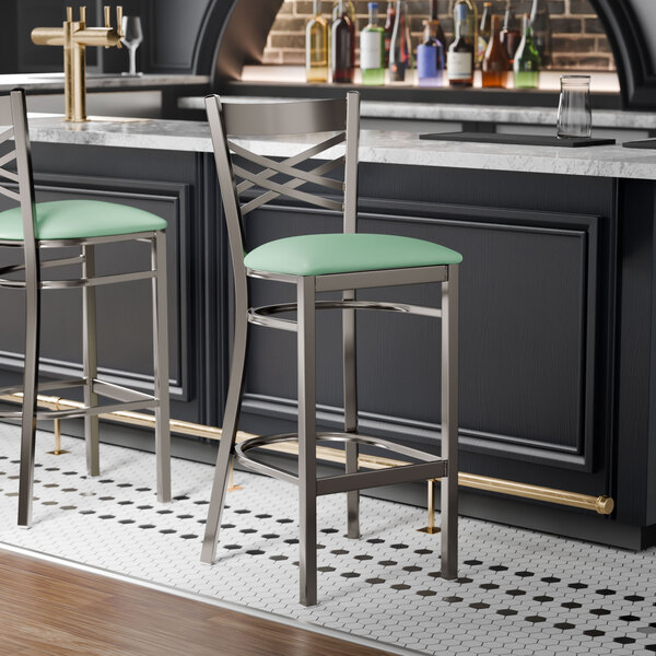 A metal bar stool with a cross back design and a seafoam green vinyl padded seat.