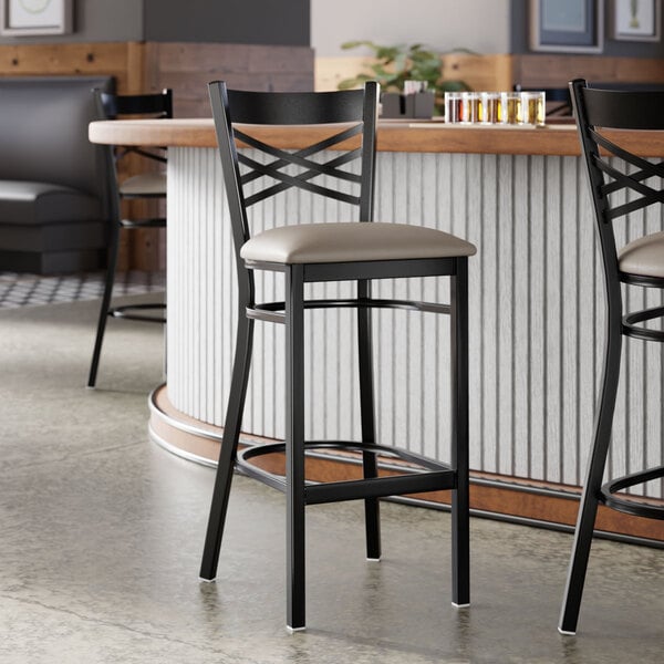 A black metal bar stool with a cross back design and a light gray vinyl padded seat.