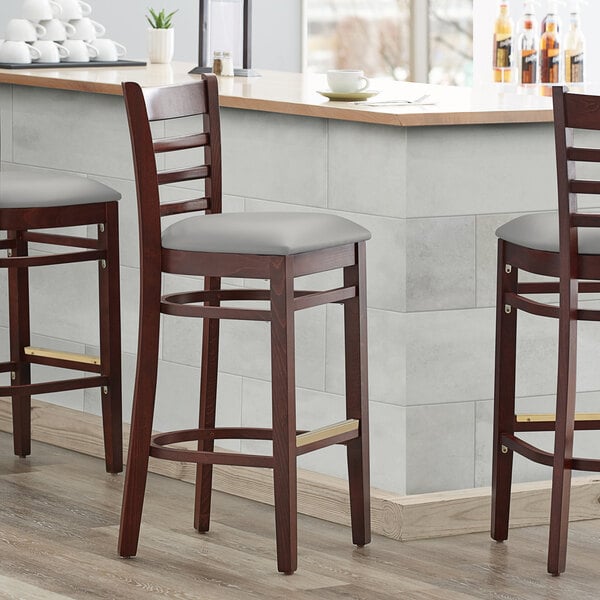 Three Lancaster Table & Seating mahogany wood bar stools with light gray vinyl seats and ladder backs at a counter.