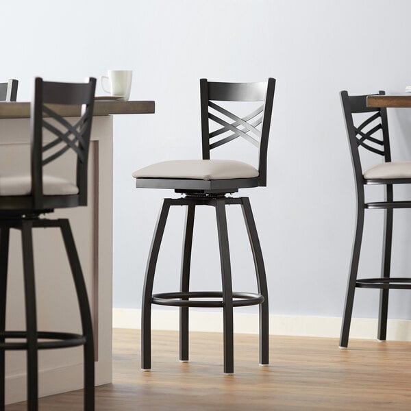 A set of three Lancaster Table & Seating black cross back swivel bar stools with light gray padded seats.