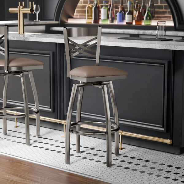 A metal bar stool with a cross back design and a taupe vinyl padded swivel seat.