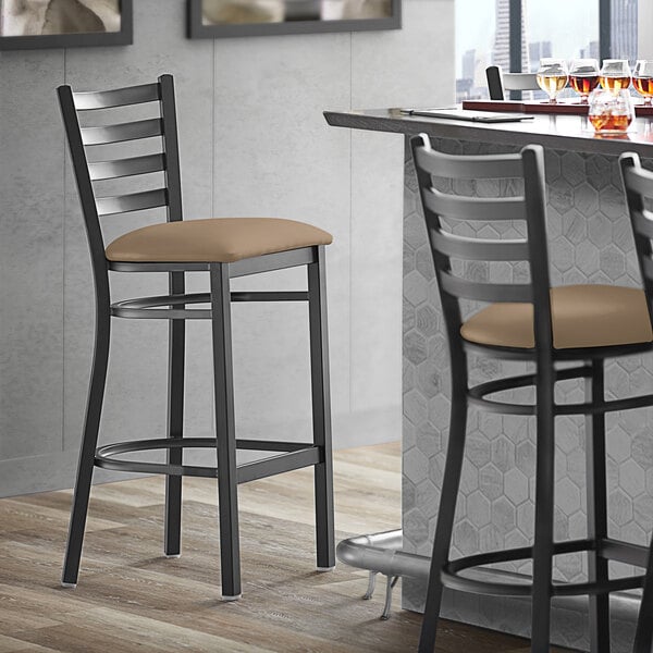 A Lancaster Table & Seating black finish ladder back bar stool with a taupe vinyl padded seat next to a table.