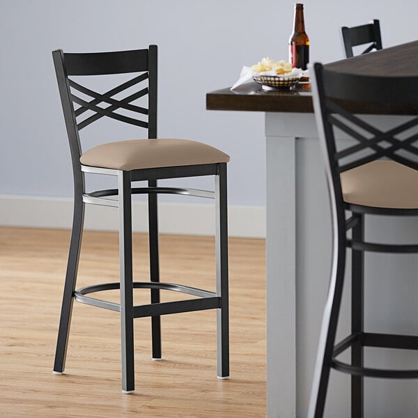 A Lancaster Table & Seating black cross back bar stool with a taupe cushion on a tan seat.