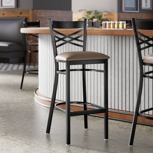 A black metal bar stool with a cross back design and a taupe vinyl padded seat.