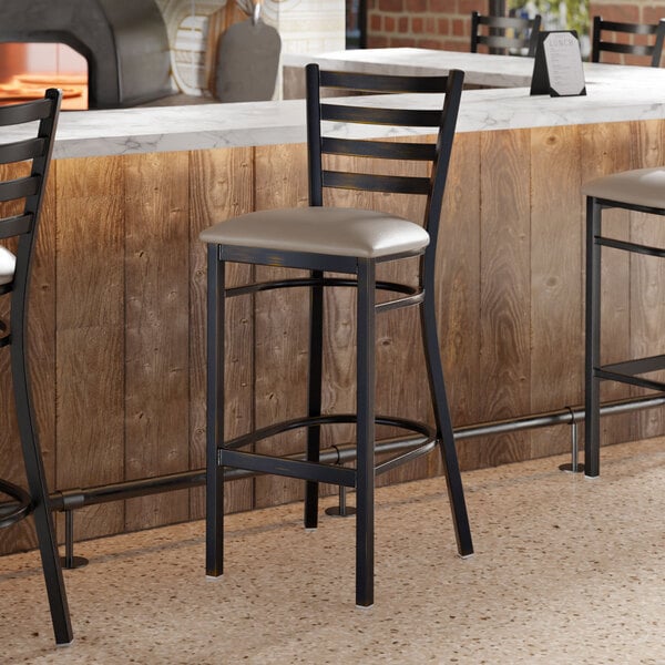 A ladder back bar stool with a distressed copper finish and a dark gray vinyl padded seat.