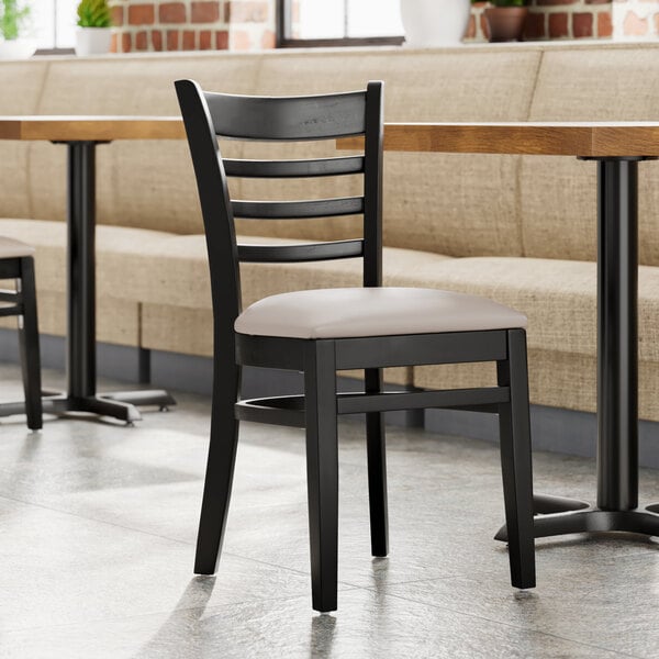 A Lancaster Table & Seating black wood ladder back chair with a light gray vinyl seat next to a table.