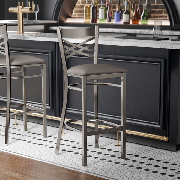 A metal bar stool with a cross back design and a dark gray vinyl padded seat.