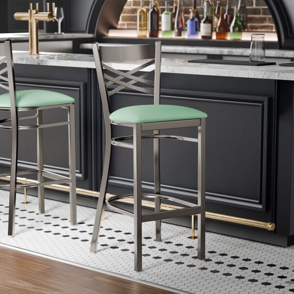 A metal bar stool with a cross back design and a seafoam green vinyl padded seat.