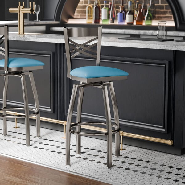 A metal bar stool with a cross back design and a blue vinyl padded seat.