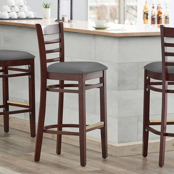 Three Lancaster Table & Seating mahogany wood ladder back bar stools with dark gray vinyl seats on a counter.