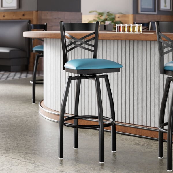 A black metal bar stool with a cross back design and a blue vinyl padded swivel seat.