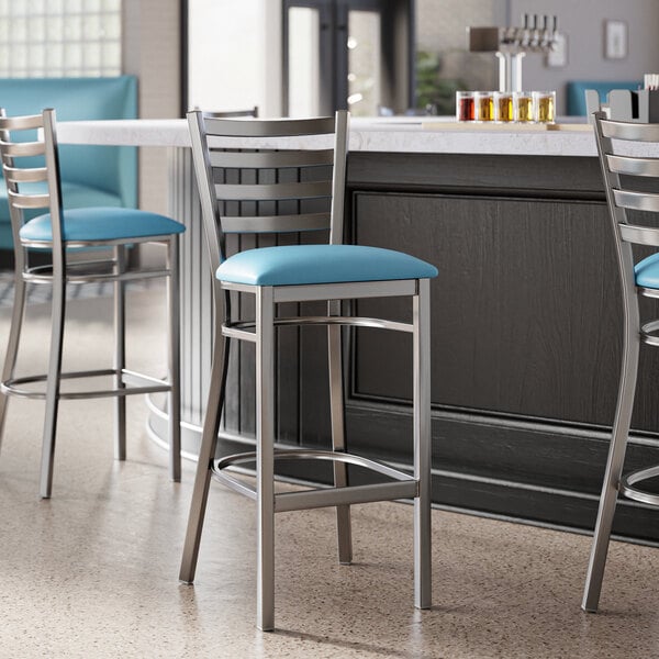 A metal bar stool with a ladder back design and a blue vinyl padded seat.