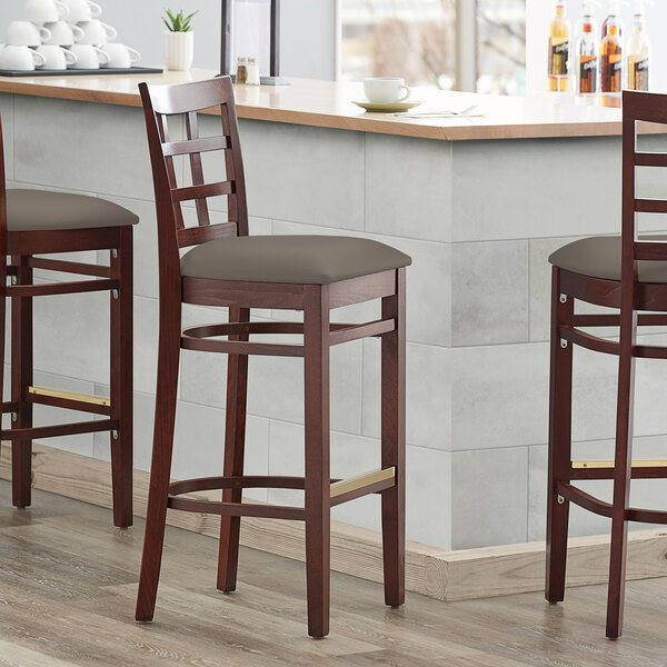 Three Lancaster Table & Seating mahogany wood bar stools with taupe vinyl cushioned seats at a bar counter.