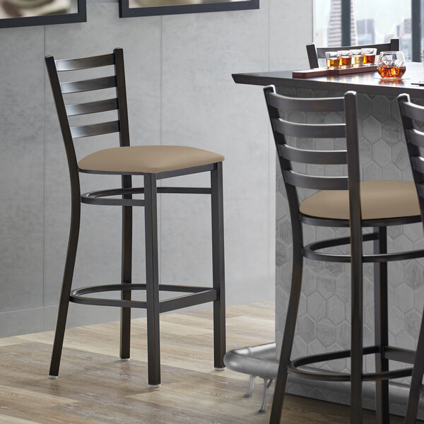 A Lancaster Table & Seating distressed copper ladder back bar stool with taupe vinyl padded seats next to a table.