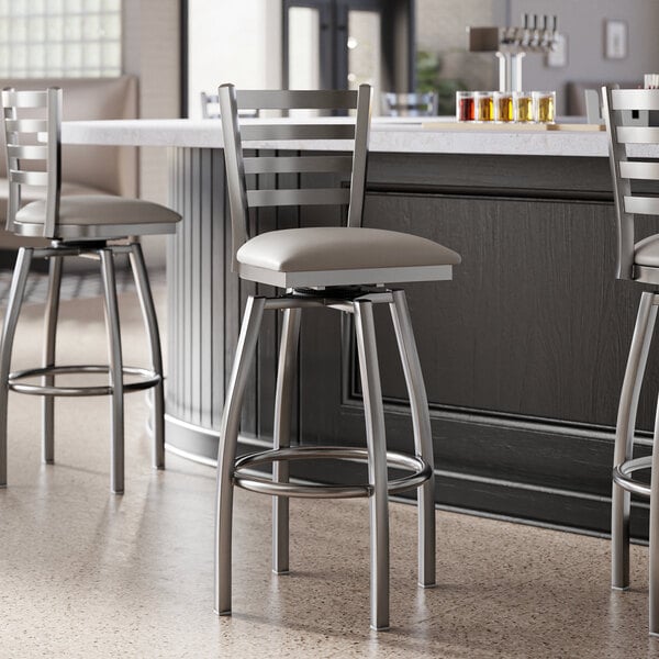 A group of Lancaster Table & Seating clear coat finish ladder back swivel bar stools with dark gray vinyl padded seats at a bar.